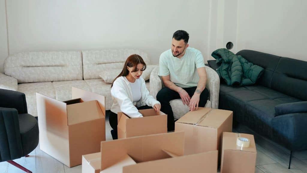 A man and a woman sit in a living room surrounded by cardboard boxes.
