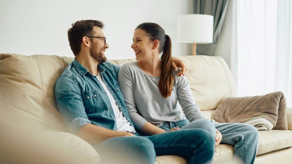 A couple sits on a couch, looking at each other and smiling while talking.