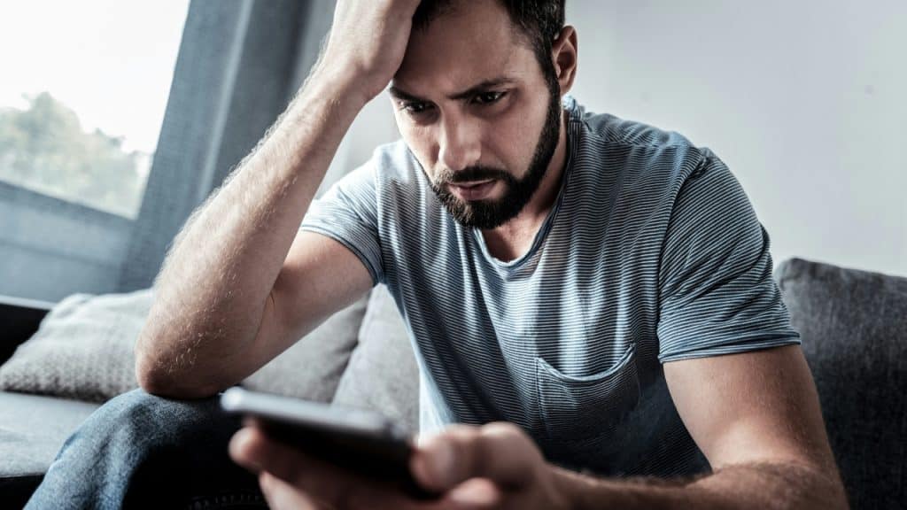 A man with a beard sits on a sofa looking upset at his phone.