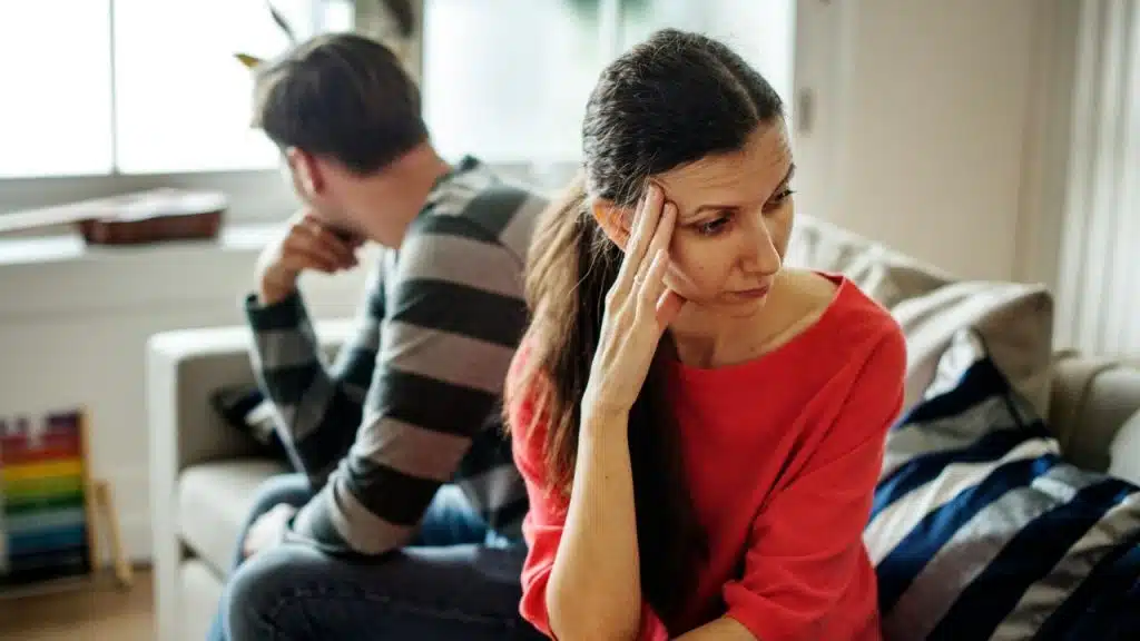A woman in a red shirt sits on a sofa touching her forehead while facing away.