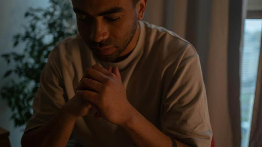 A man sitting with clasped hands in quiet reflection indoors.