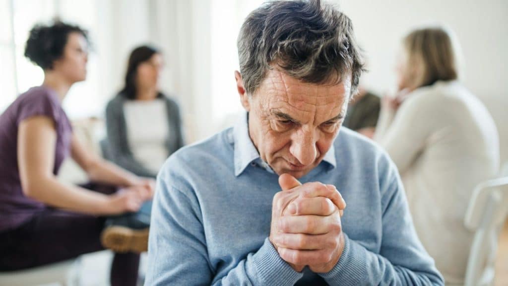An older man in a blue sweater sits with his hands clasped, looking down thoughtfully.