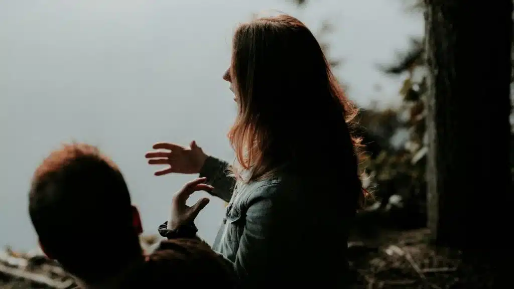 Woman gesturing with her hands while talking to a man outdoors by a foggy lake.