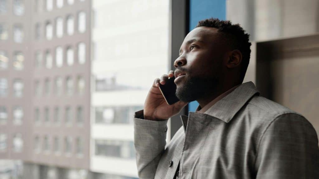 A man in a gray coat talking on a phone near a window overlooking city buildings.