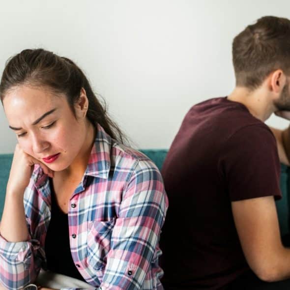 A man and woman sit back-to-back on a green sofa, looking away with downcast expressions.