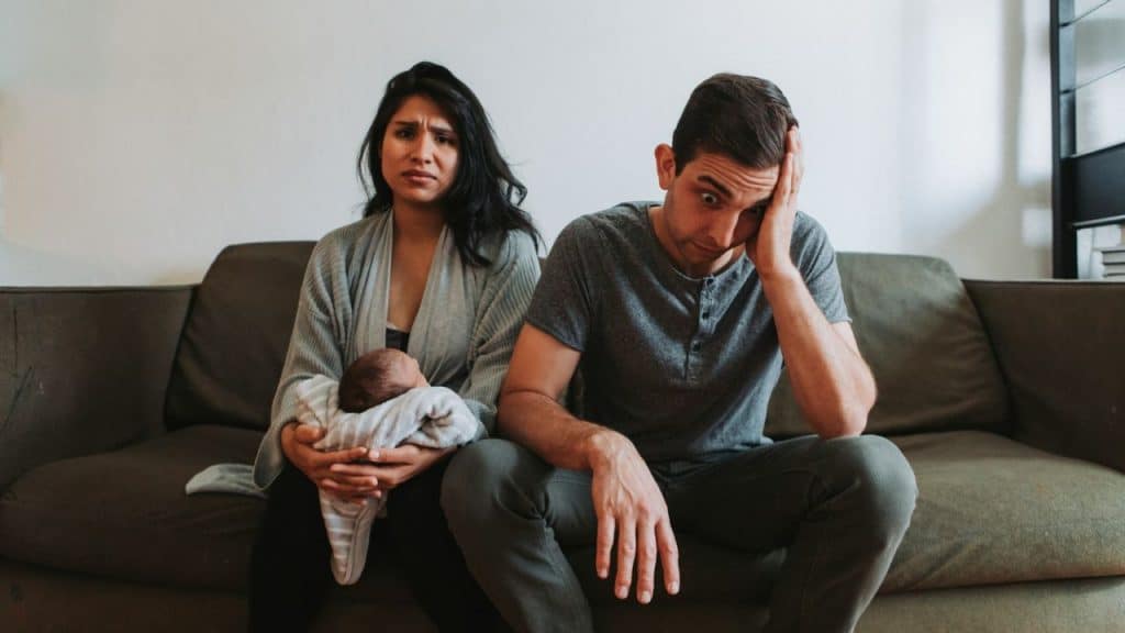A woman holding a newborn baby sits next to a stressed, weary-looking man.