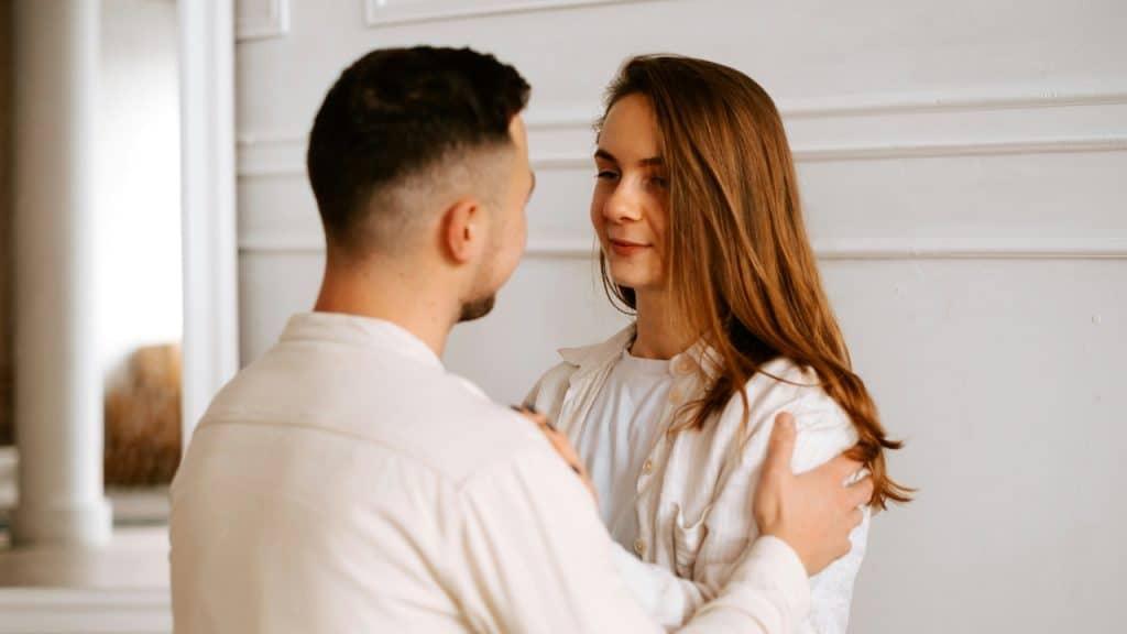 A man places his hands on a woman's shoulders as they look at each other.