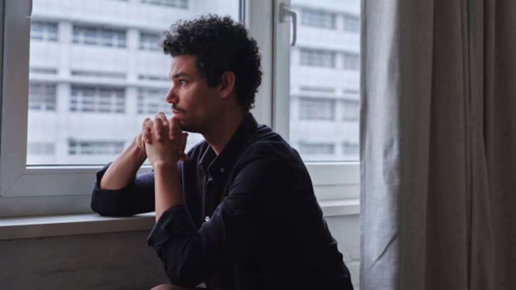 A man with curly hair sits by a window with his hands clasped together.