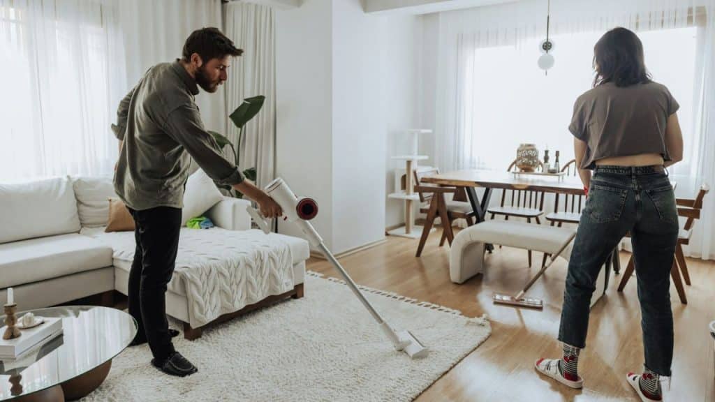 A man vacuums a white rug while a woman mops the floor in a living room
