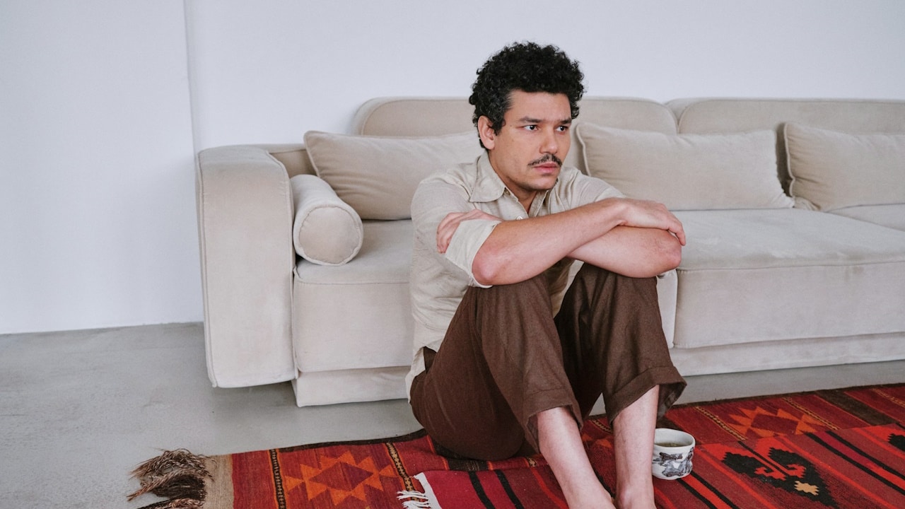A mustached man sitting with his arms folded on his knees on a carpet on the floor.