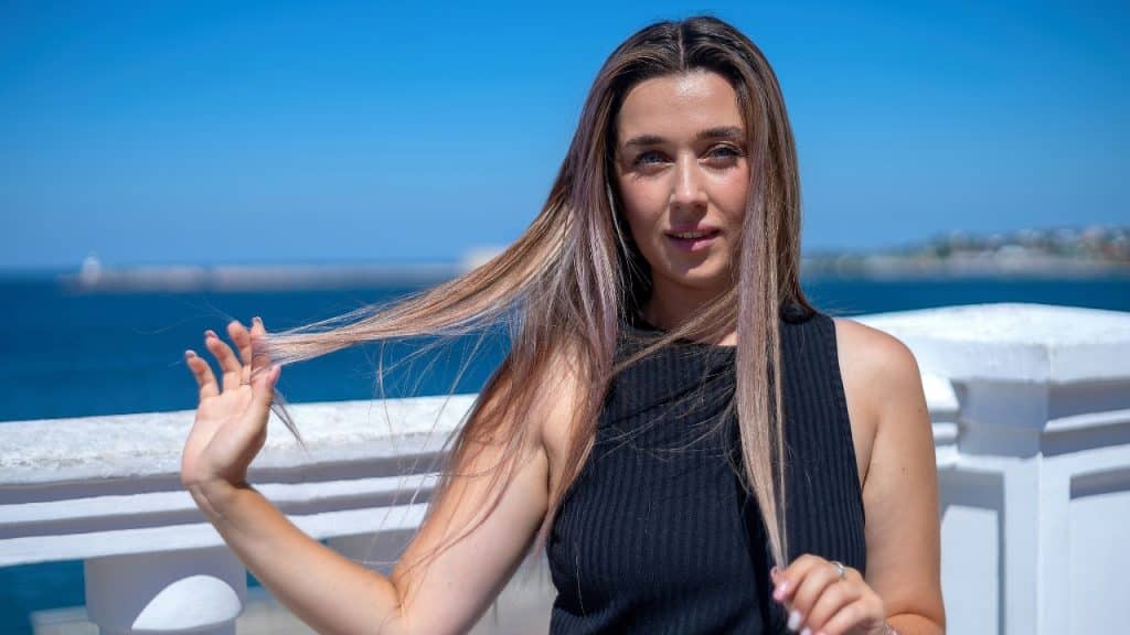 A woman with long hair stands in front of a blue ocean and white railing.