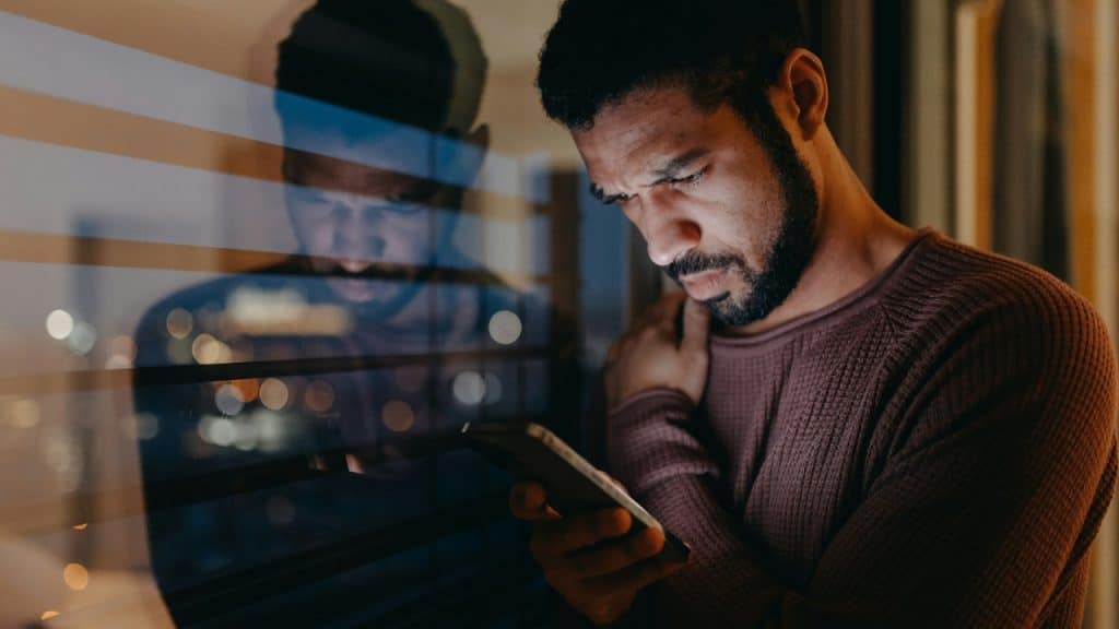 A man looks down at his phone at night with his reflection visible in glass.