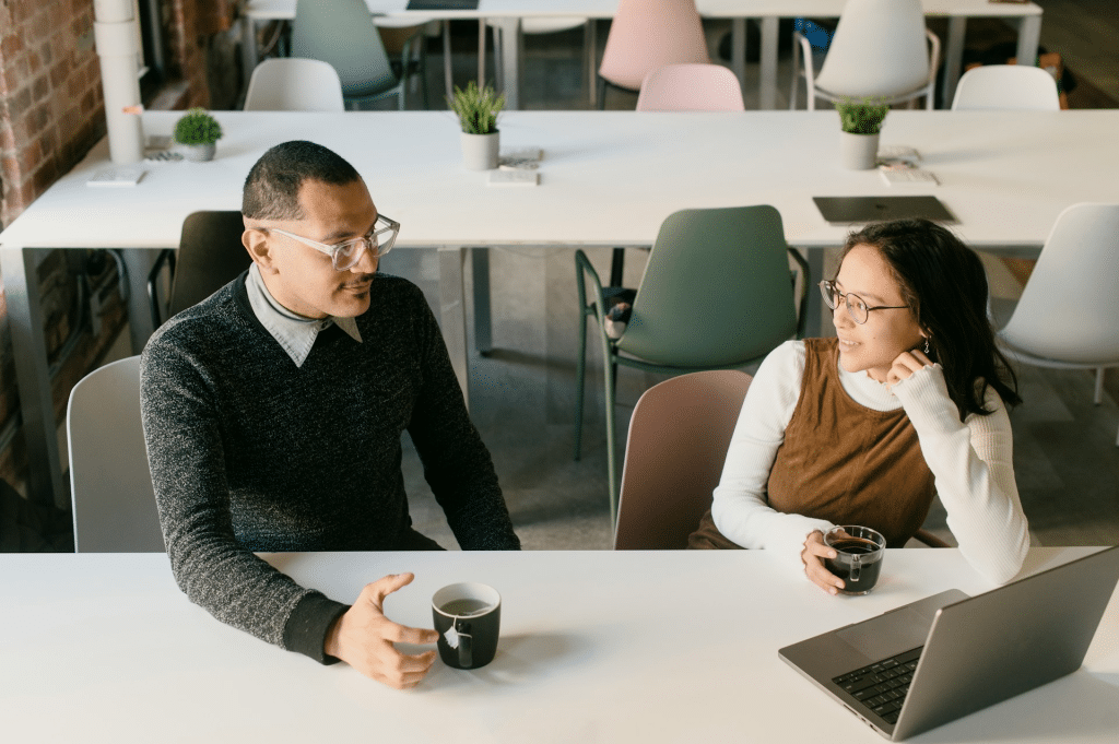 Two people wearing glasses are sitting at a white table, talking and drinking coffee with a laptop present in a modern, open-office setting.