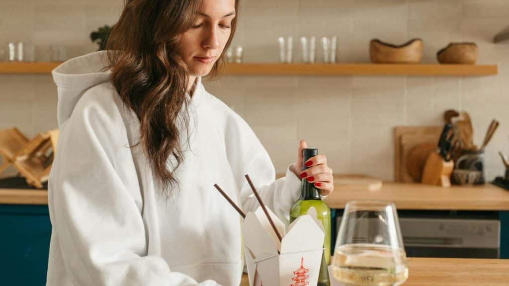 A woman opening a bottle beside a takeout container and wine glass.