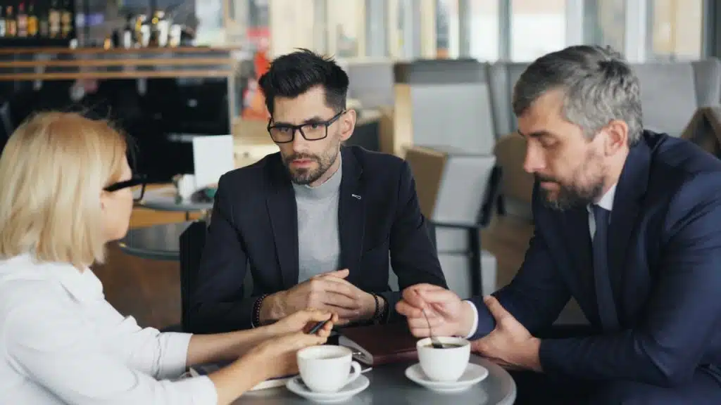 A woman and two men in business attire talk around a small café table.