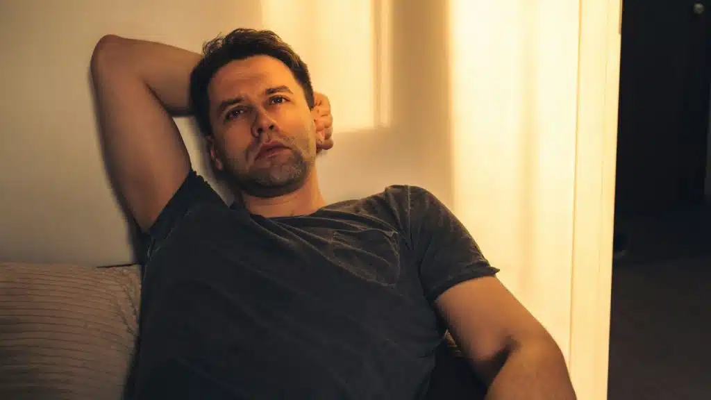 A man in a dark t-shirt leans against a wall in warm, golden indoor lighting.