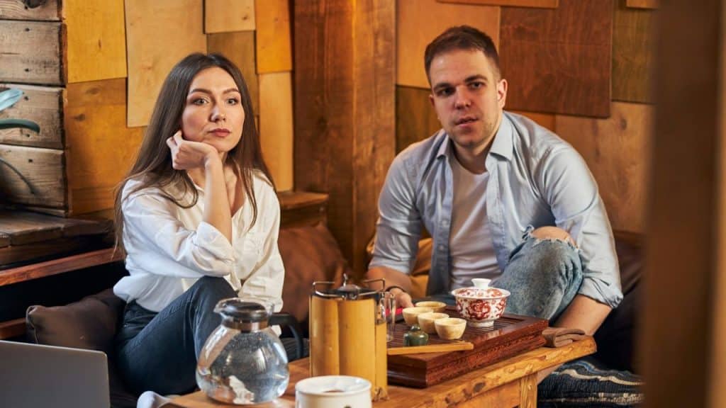 A man and woman sit at a low table with tea sets in a wood-paneled room.