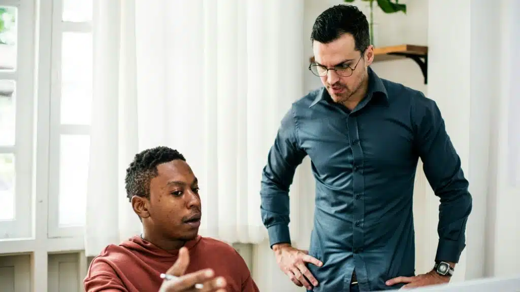 A man in a blue shirt stands over a seated man, gesturing with focused expression.