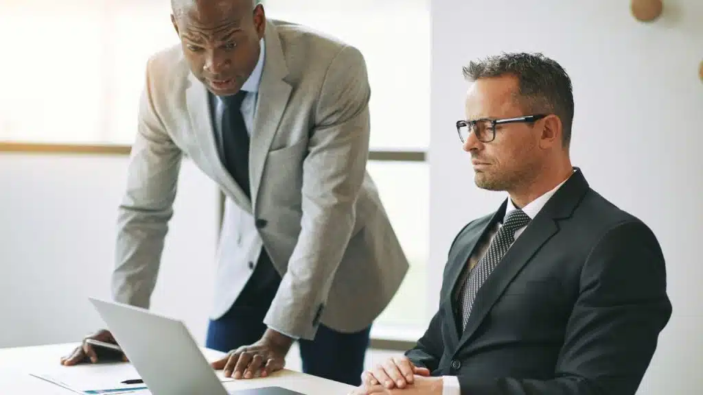 Two men in suits look at a laptop with serious expressions in a bright office.
