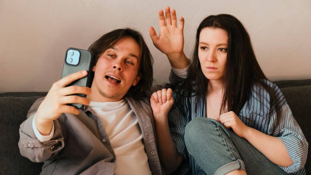 A man waving hands while making a video call and his wife is looking at the screen, seems annoyed.