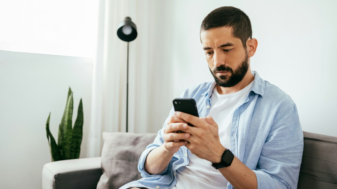 A bearded man in a light blue shirt sits on a sofa looking down at a smartphone.