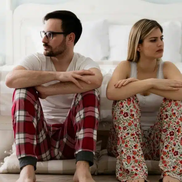 A man and woman sit on the floor in pajamas, leaning away from each other.