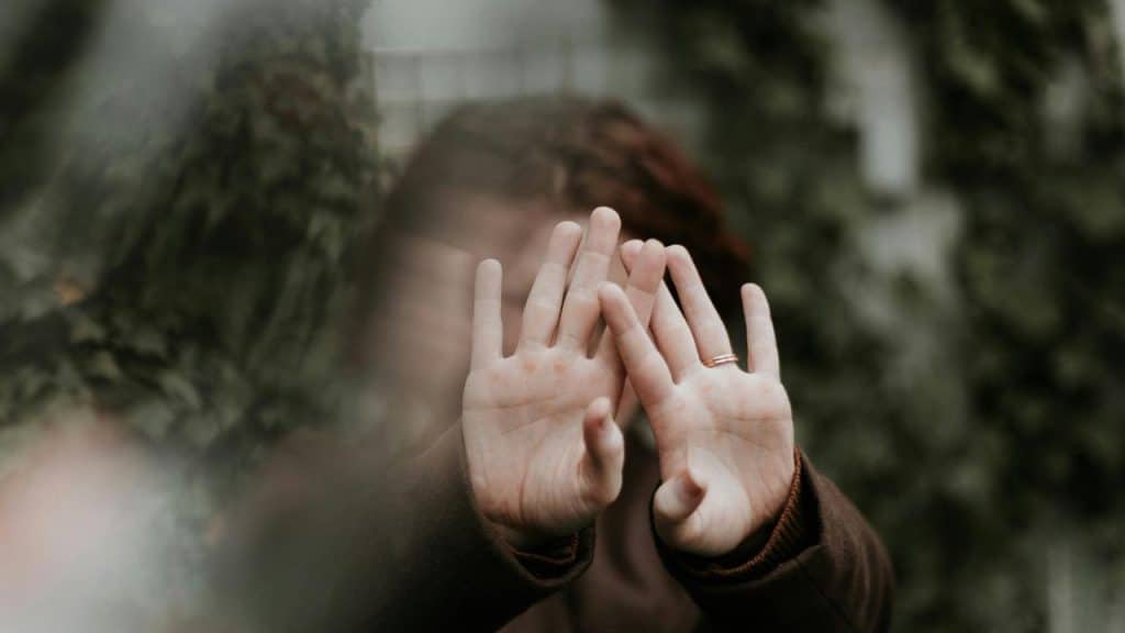 A person holding up their hands to cover their face behind glass.
