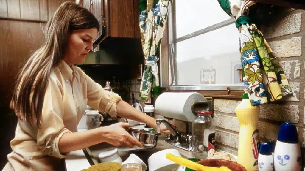 A woman washes a metal pot in a kitchen sink surrounded by various cleaning supplies.