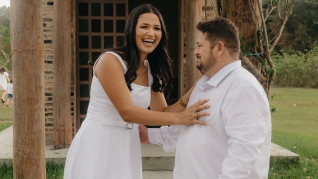 A laughing woman in a white dress places her hand on a man's chest outdoors.