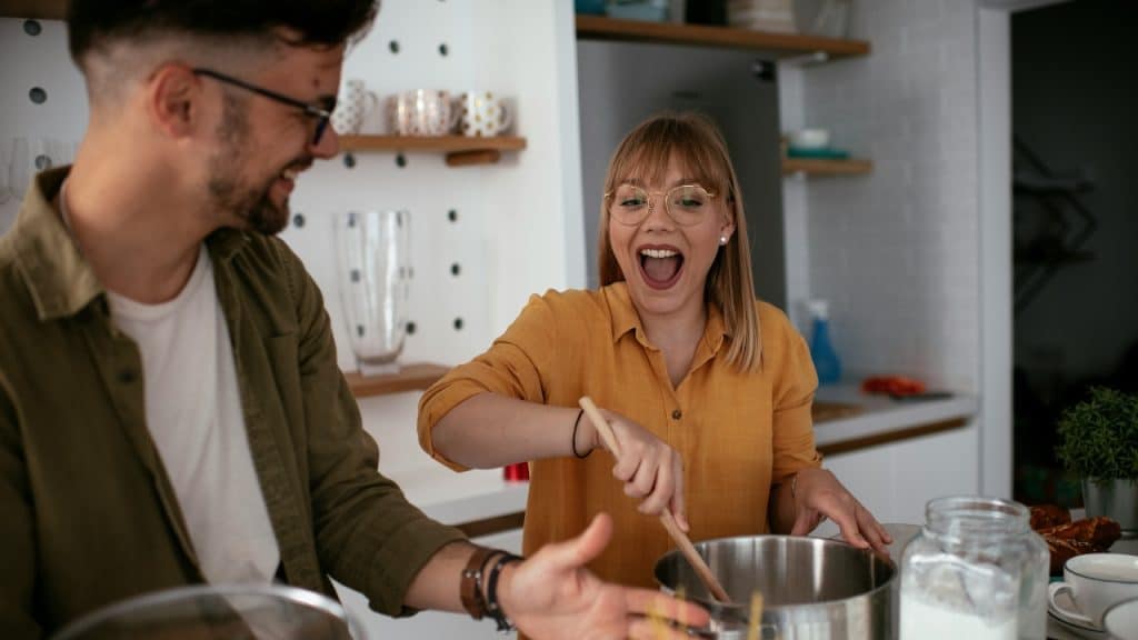 A smiling woman stirs a metal pot with a wooden spoon while a man looks on nearby.
