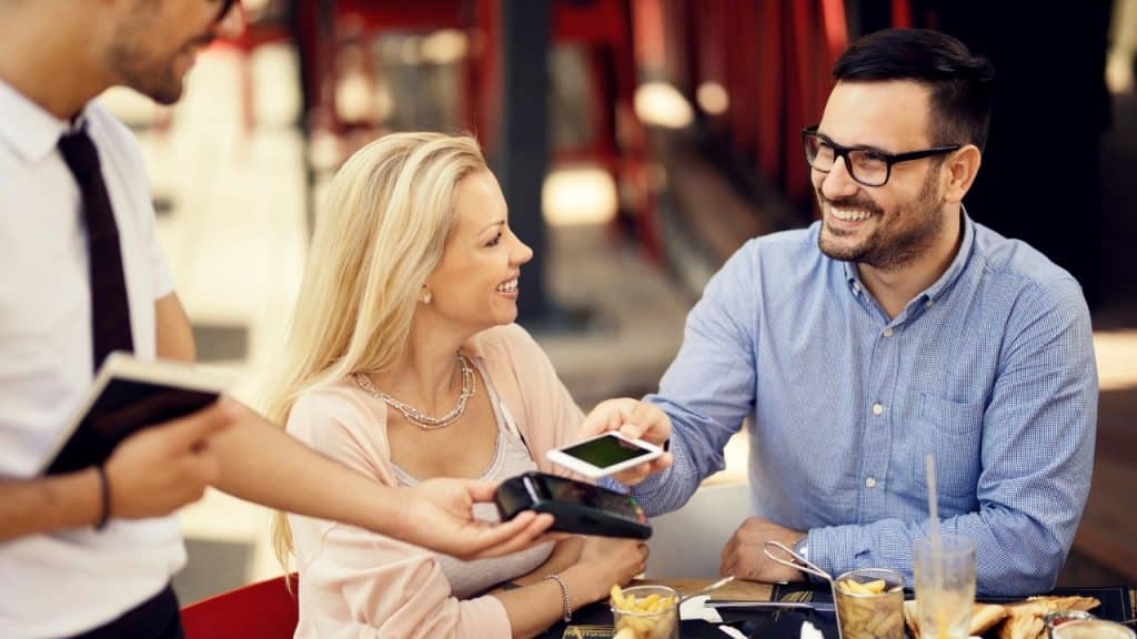 A smiling man uses a smartphone to pay at a handheld terminal held by a waiter.