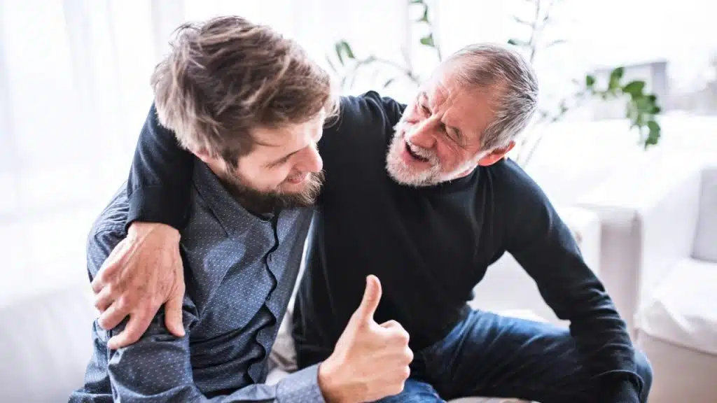An older man with a grey beard smiles and gives a thumbs up to a younger man.