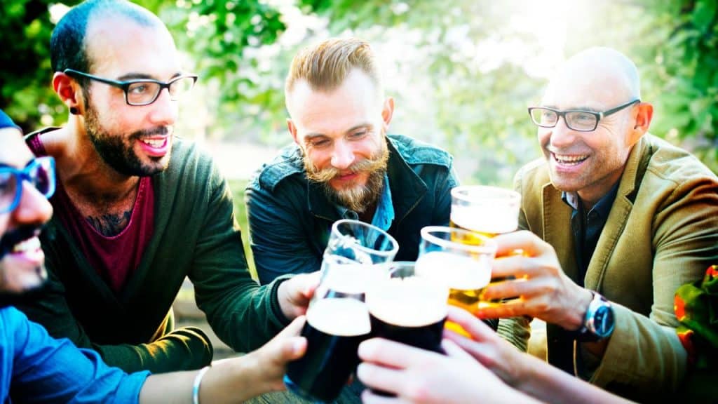 Four men outdoors sitting at a table together while clinking several glasses of dark beer.