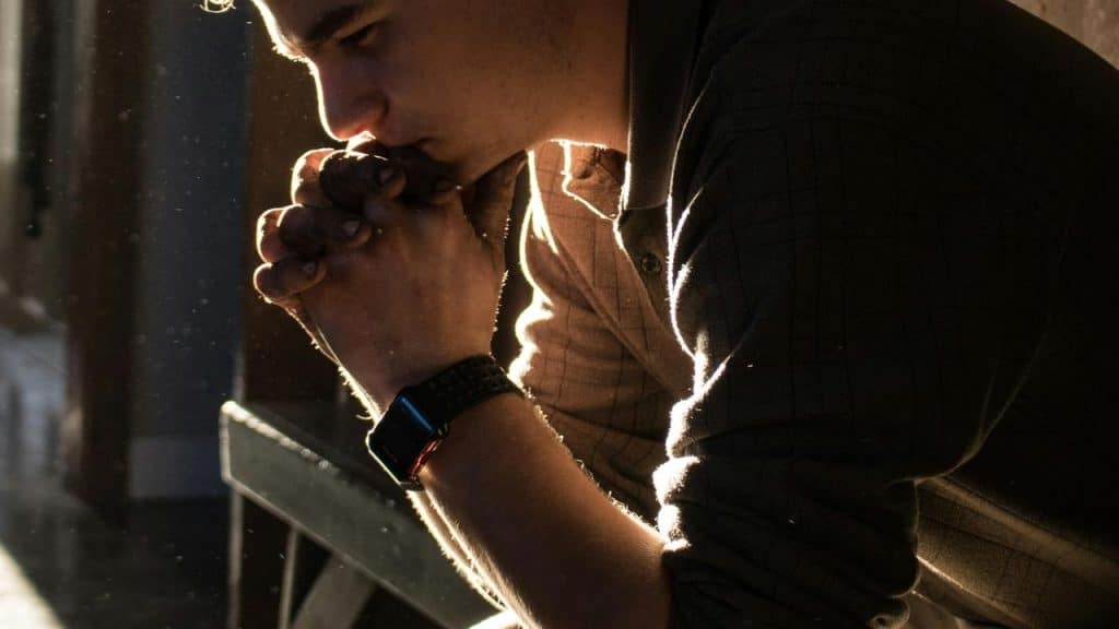 A man sitting in warm sunlight, leaning forward with hands clasped in thought.