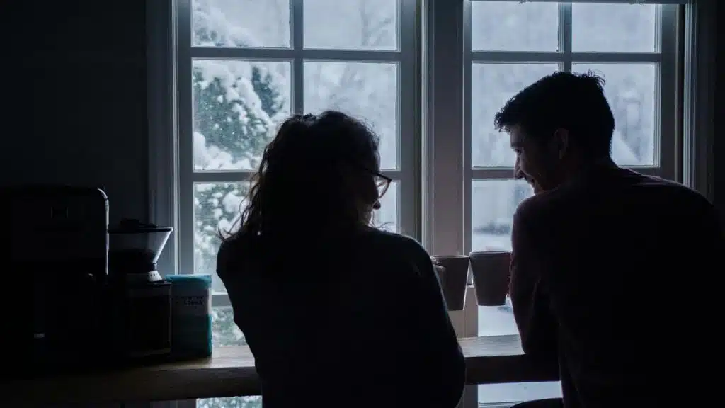 A couple sitting by a window, sharing coffee and smiling on a snowy day.