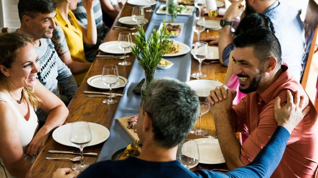 A group of people sits around a long wooden dining table talking and laughing together.