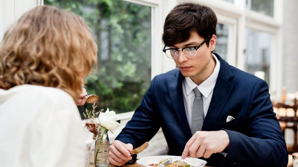 A man in a blue suit and glasses looks down at his meal during a silent dinner.
