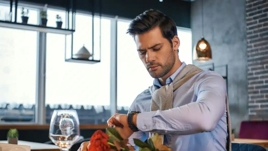 A man in a blue shirt sits at a table and looks at his wristwatch.