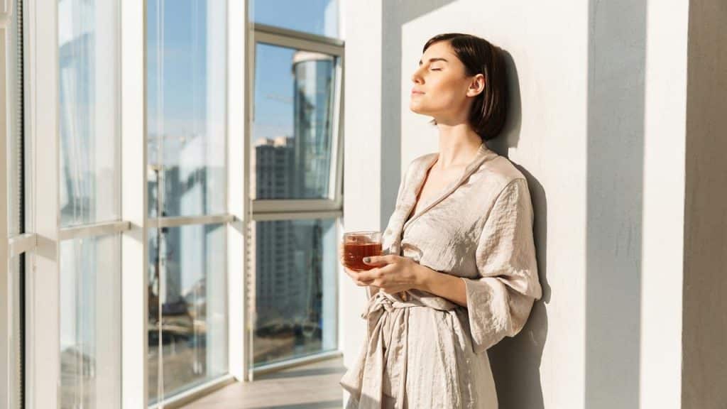 A woman standing by a window with her eyes closed holding a cup of tea.