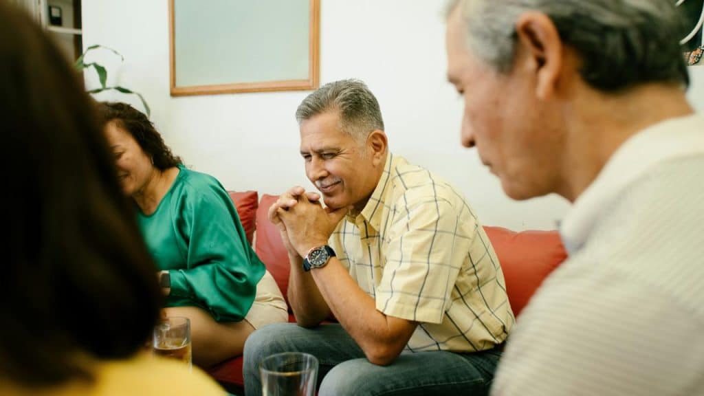 A group of adults sit together in a living room, appearing thoughtful and engaged.