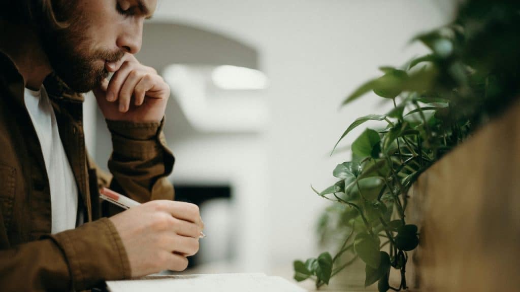 A man with a beard leans over a desk while writing in a notebook near plants.