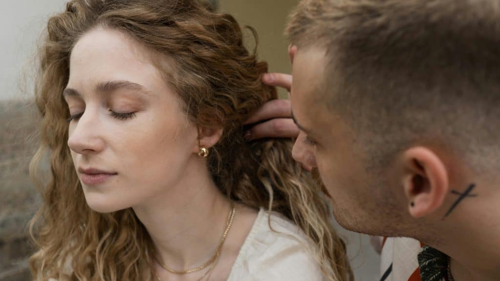 A man with a cross tattoo on his neck touches a woman's long curly hair.