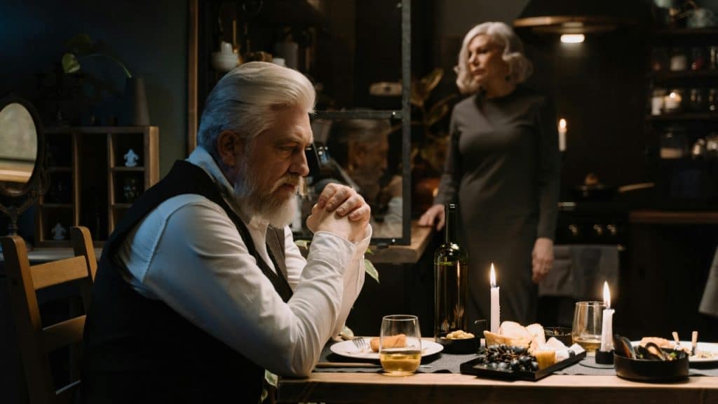 An older man sits at a candlelit dinner table while a woman stands behind him.