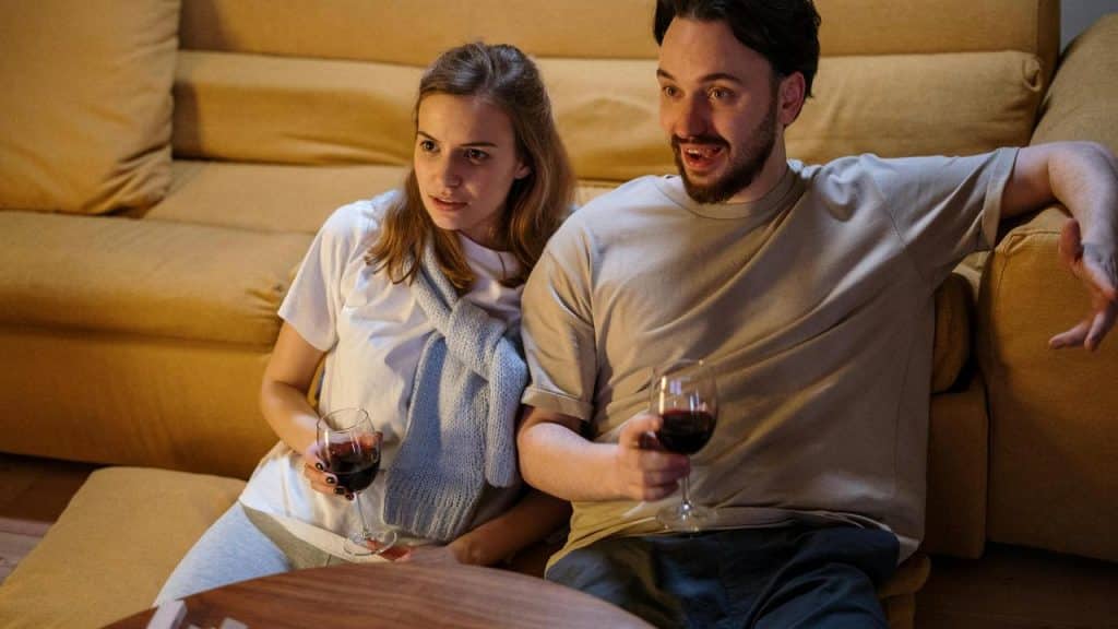 A couple sitting on a couch holding glasses of wine and watching something.