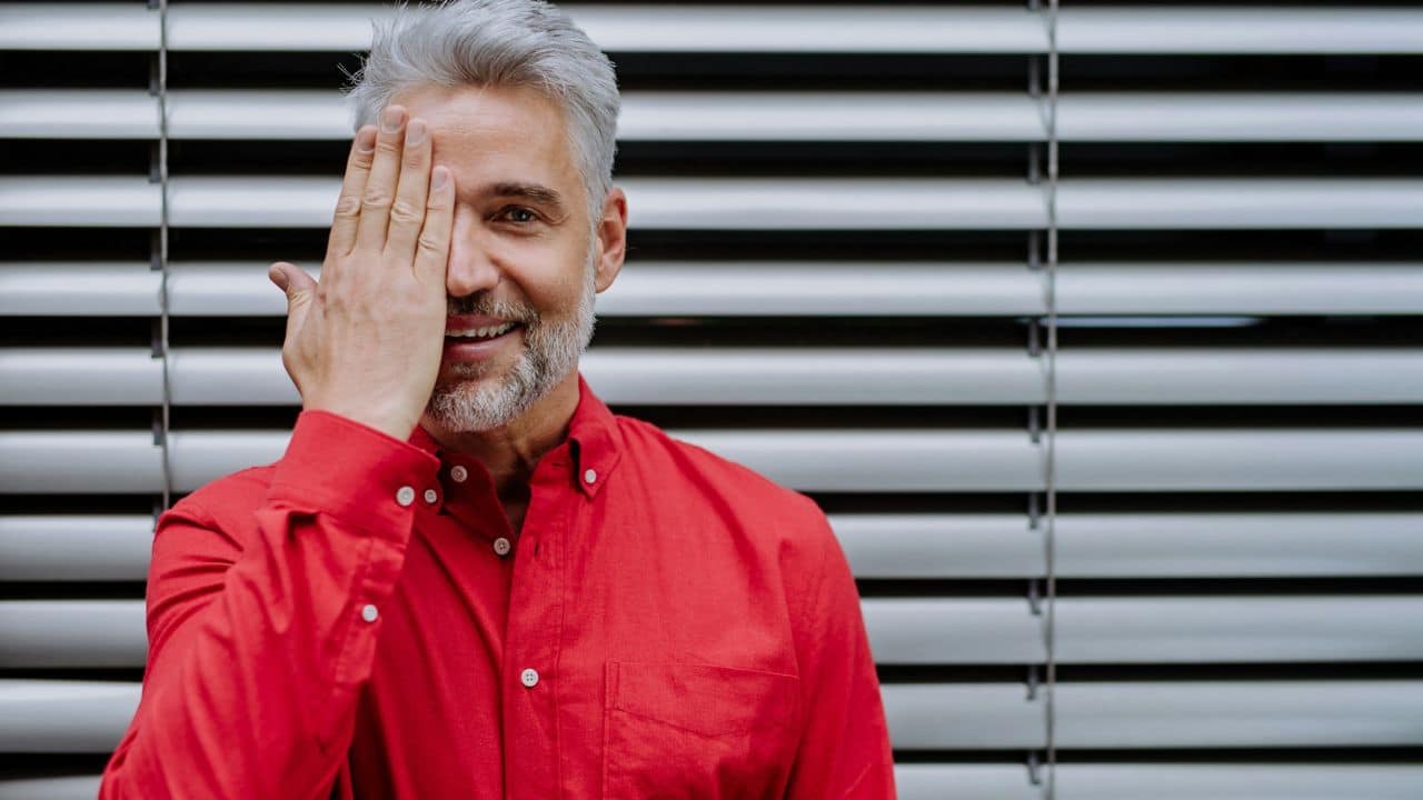 A smiling man with gray hair covers one eye with his hand against slatted blinds.
