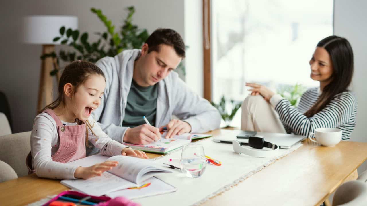 A man and young girl write in books while a woman sits nearby and smiles.
