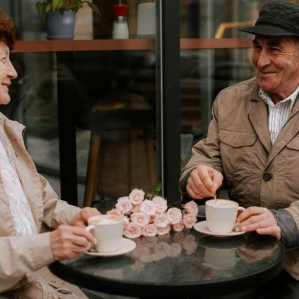 An elderly couple smiles at each other while having coffee at an outdoor table.