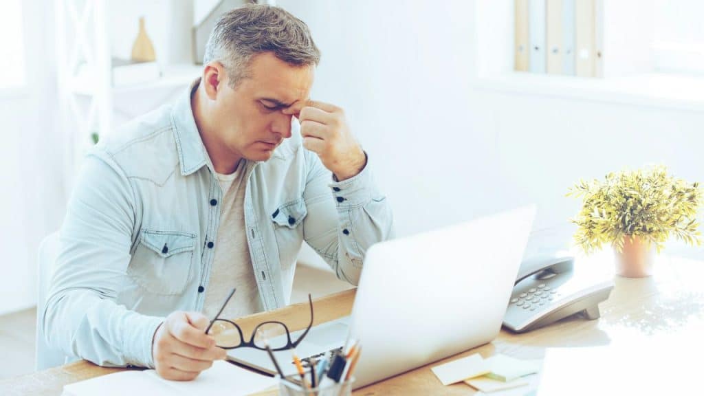 A man at a desk holds glasses and pinches the bridge of his nose near a laptop.
