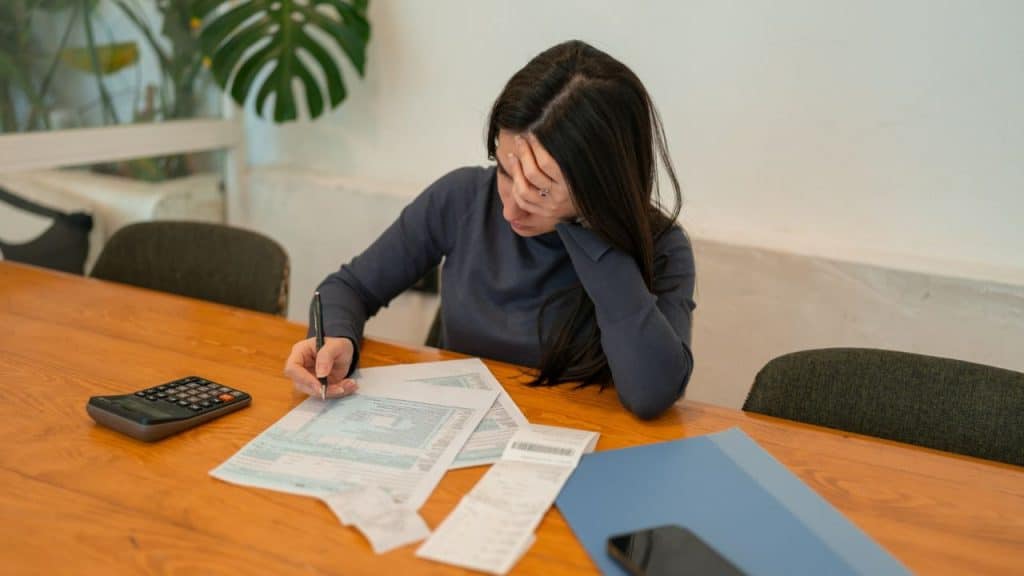 A woman sits at a table with a calculator, writing on papers with a stressed expression.