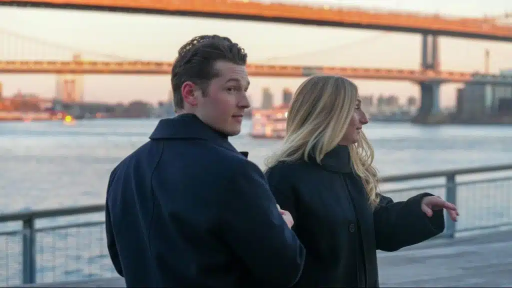 A couple standing by a riverside railing with city bridges in the background.